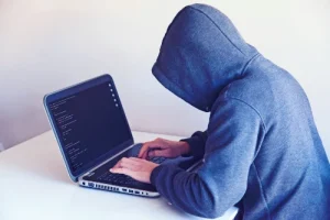 A person hidden in a grey hoodie is usineg a laptop computer. It is assumed that this person is a hacker.